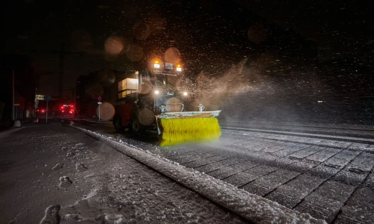 Snerydningskøretøj med gul roterende kost fjerner sne fra byvej om natten under snevejr.