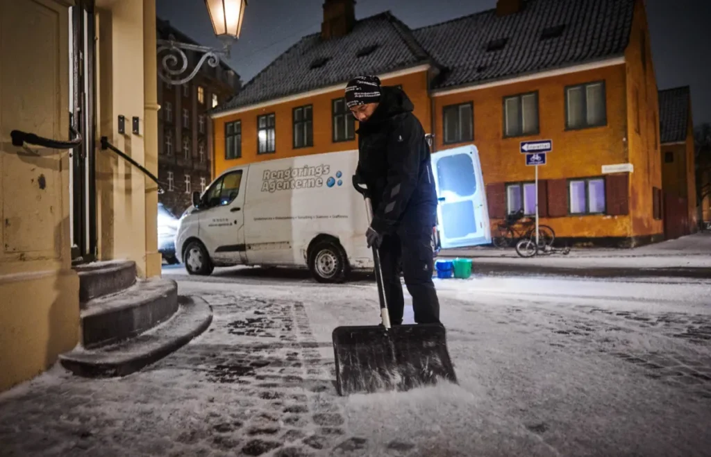 Medarbejder skovler sne foran bygning om natten.